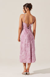 A woman is wearing a pink dress with a subtle floral pattern. The dress has thin straps, a low-cut back with crisscross lacing, and ties at the waist. The length of the dress reaches mid-calf. She is also wearing light-colored heeled shoes.
